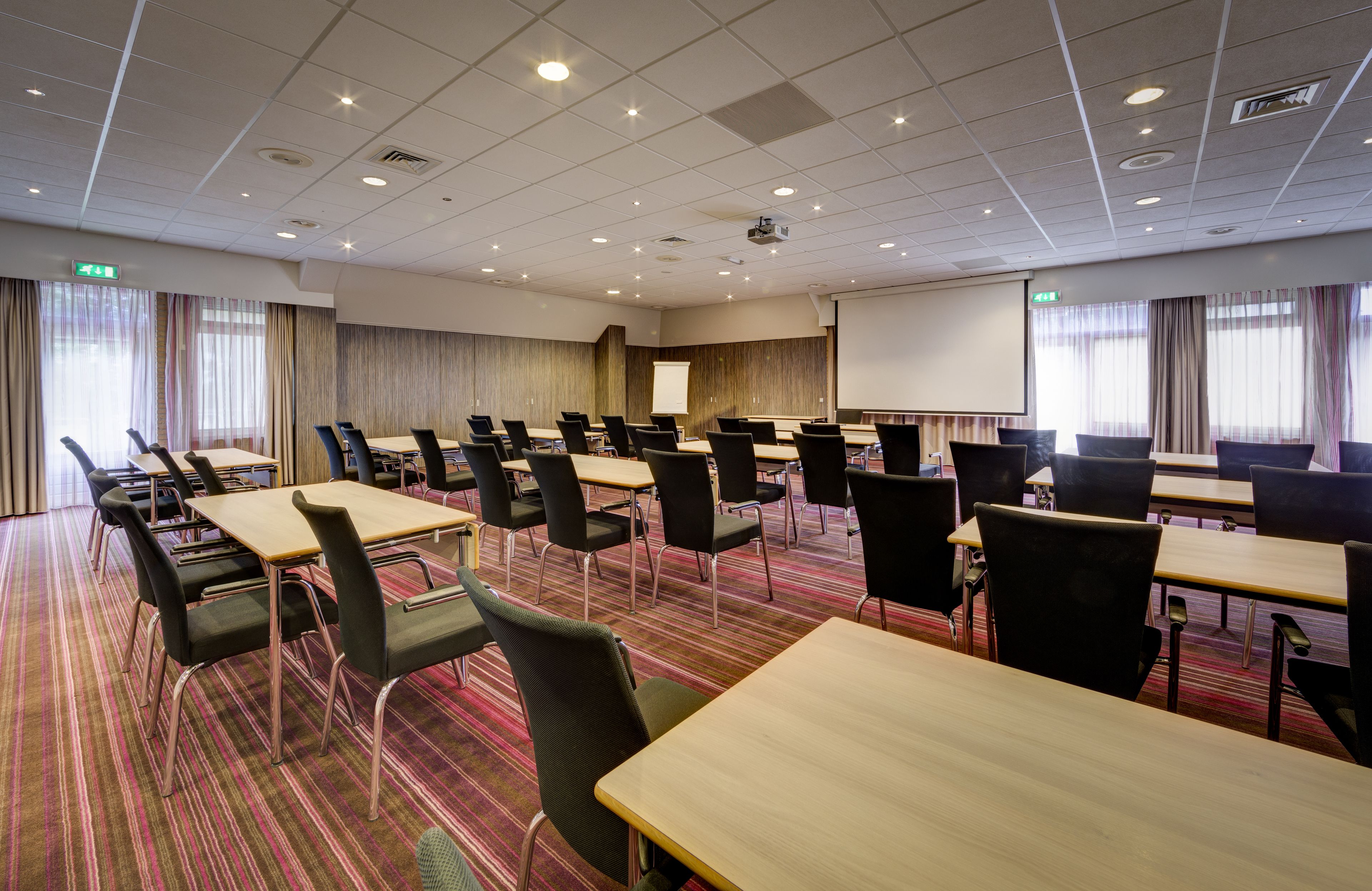 A conference room with multiple tables and chairs, a projector screen, and a large window with curtains.