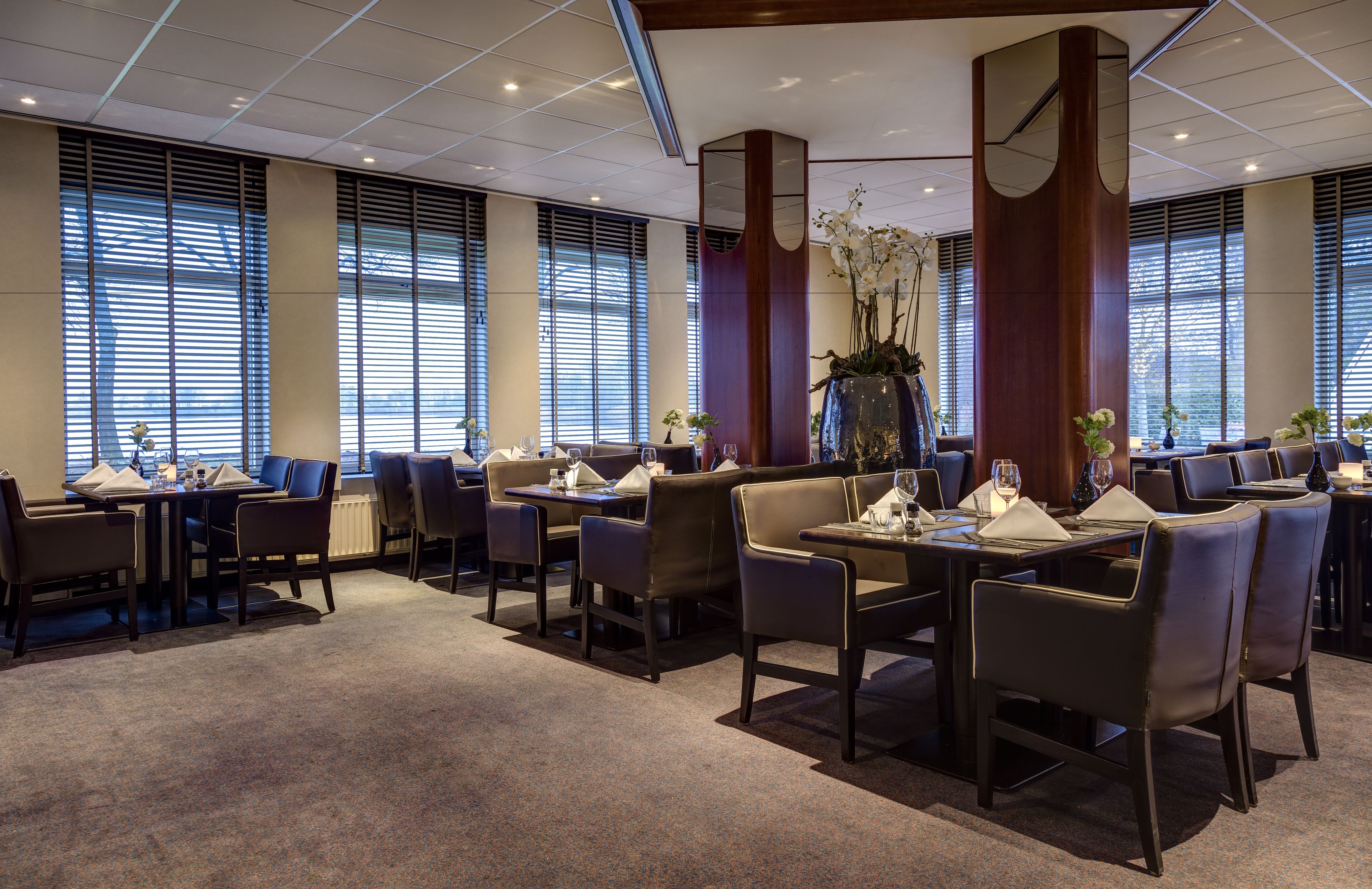 A modern dining area with multiple tables and chairs, large windows with blinds, and decorative plants.