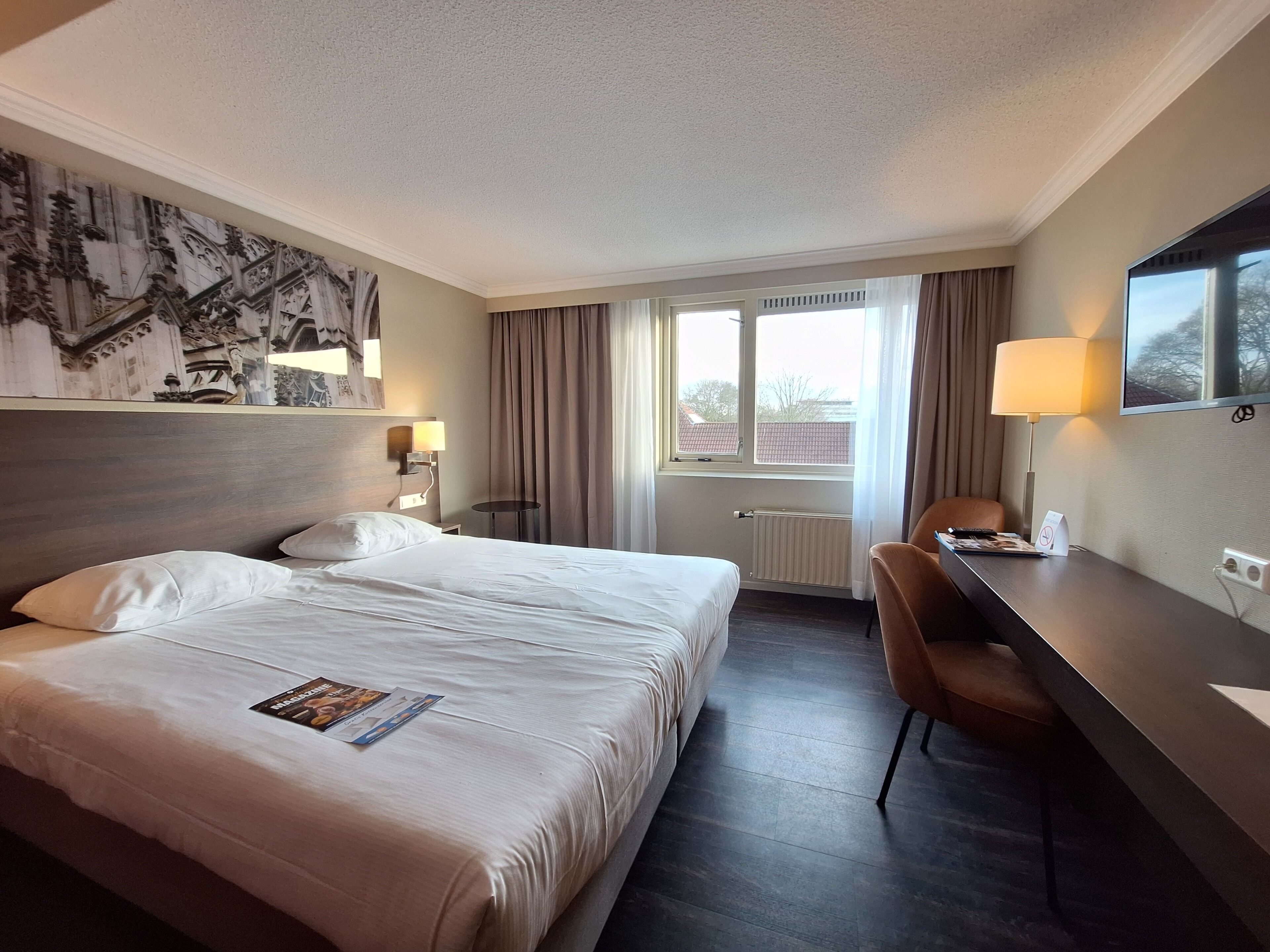 Habitación de hotel con una cama grande, un escritorio con silla, un televisor y una ventana con cortinas.