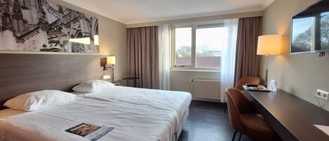 Een hotelkamer met een groot bed, een bureau met stoel, een televisie en een raam met gordijnen.