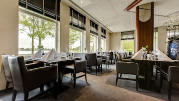 Un comedor moderno con varias mesas y sillas, ventanas amplias con persianas y vistas a árboles.