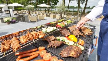 Una barbacoa al aire libre con diversos tipos de carne y verduras a la parrilla, rodeada de mobiliario para exteriores y sombrillas.