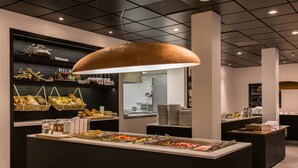 A buffet area with various food options, a large hanging light fixture, and a kitchen visible in the background. - Postillion Hotel Deventer (Deventer)