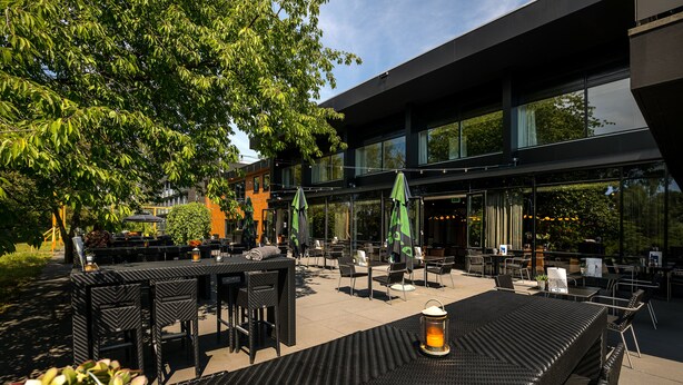 An outdoor seating area with tables and chairs, surrounded by greenery and a modern building with large windows. - Postillion Hotel Deventer (Deventer)