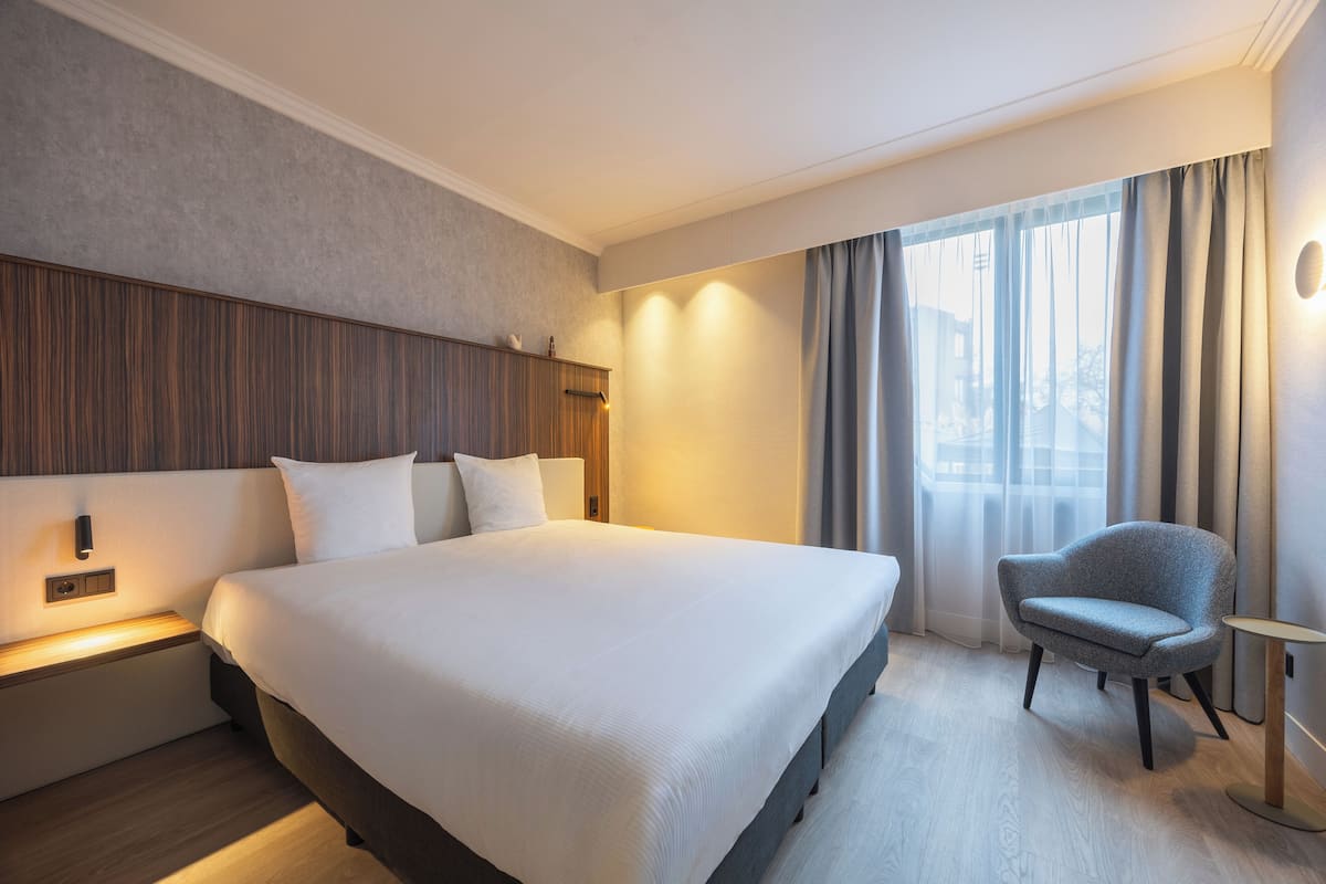 A modern hotel room with a large bed, a nightstand, a chair, and a window with curtains.