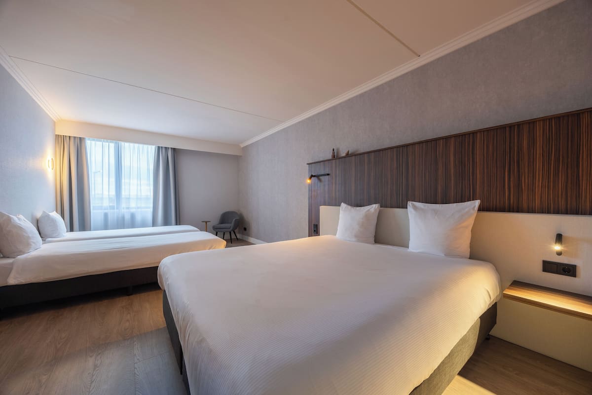 A hotel room with two beds, a wooden floor, a large window with curtains, a bedside table with a lamp, and a wall-mounted shelf.
