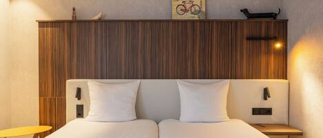 Cama doble con sábanas blancas, cabecera de madera, mesita de noche pequeña con lámpara y teléfono instalado en la pared.