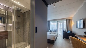 A modern hotel room with a glass-enclosed shower, a bed with a view of the city, a desk with a chair, and a flat-screen TV on the wall. - Postillion Hotel Utrecht Bunnik (Bunnik)