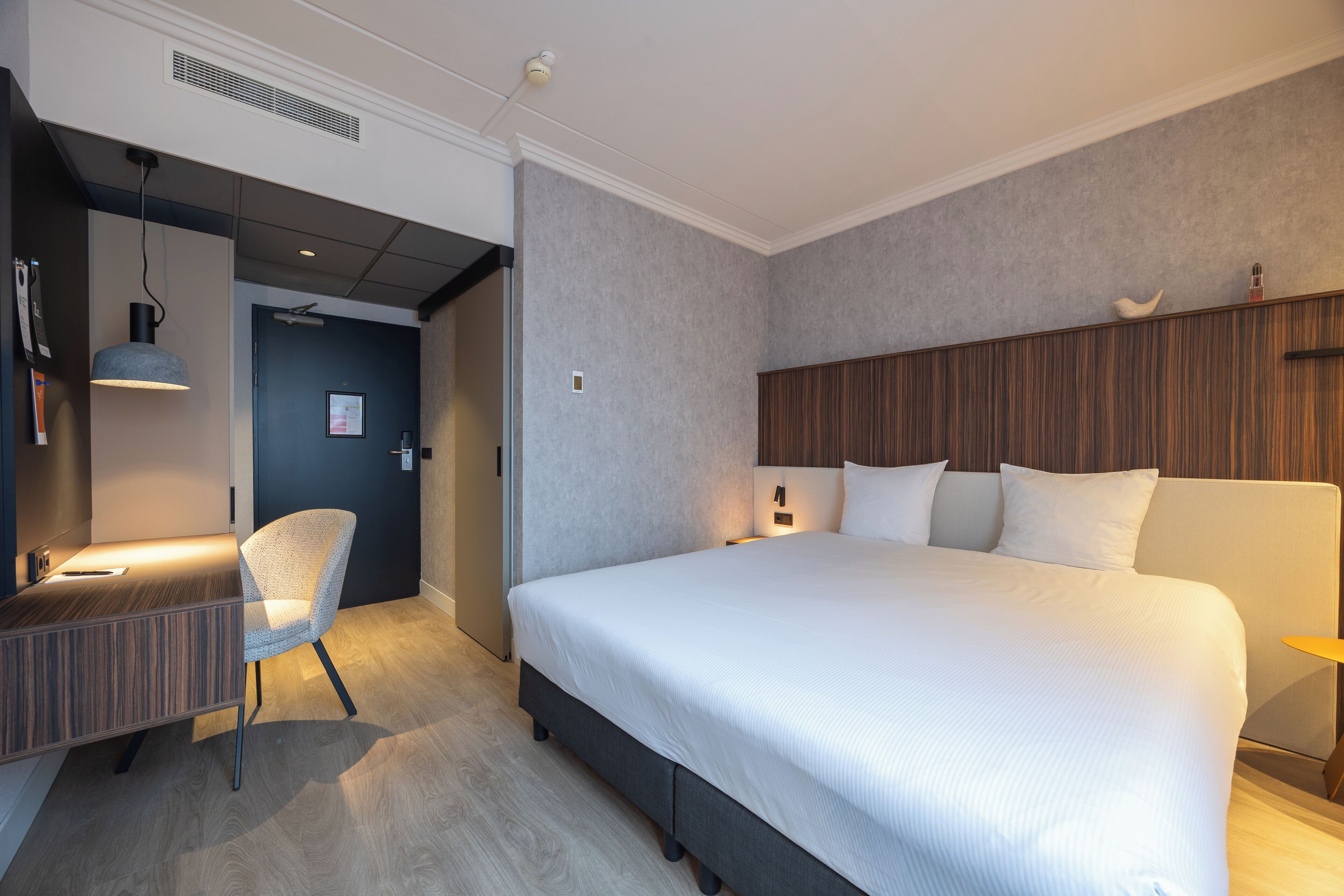 Ein modernes Hotelzimmer mit einem großen Bett, einem Schreibtisch mit Stuhl und einem kleinen Tisch mit Lampe.