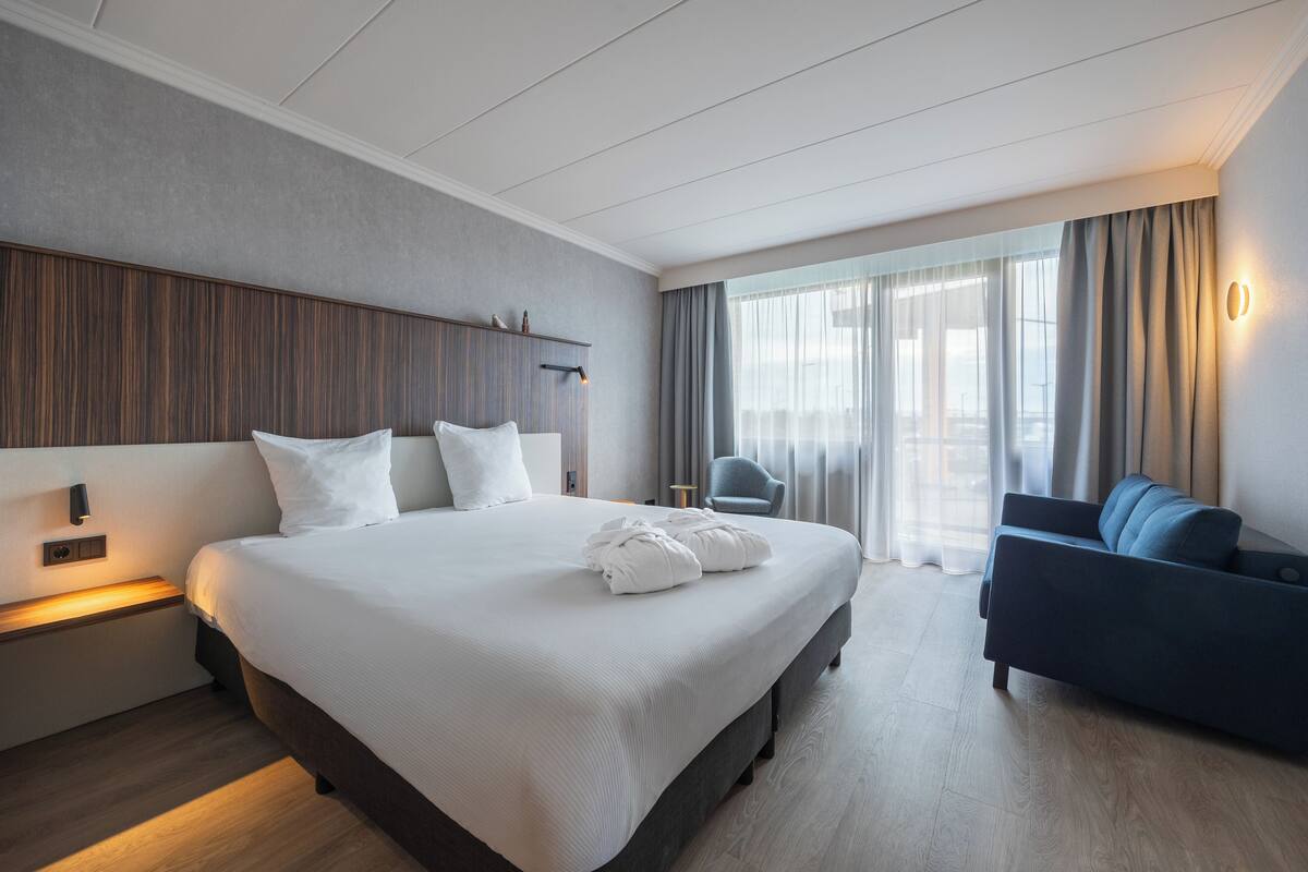A modern hotel room with a large bed, a sofa, a desk, and a window with curtains.