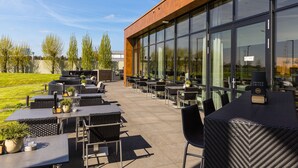 An outdoor patio area with tables and chairs, a wooden building facade, and a grassy lawn. - Postillion Hotel Utrecht Bunnik (Bunnik)