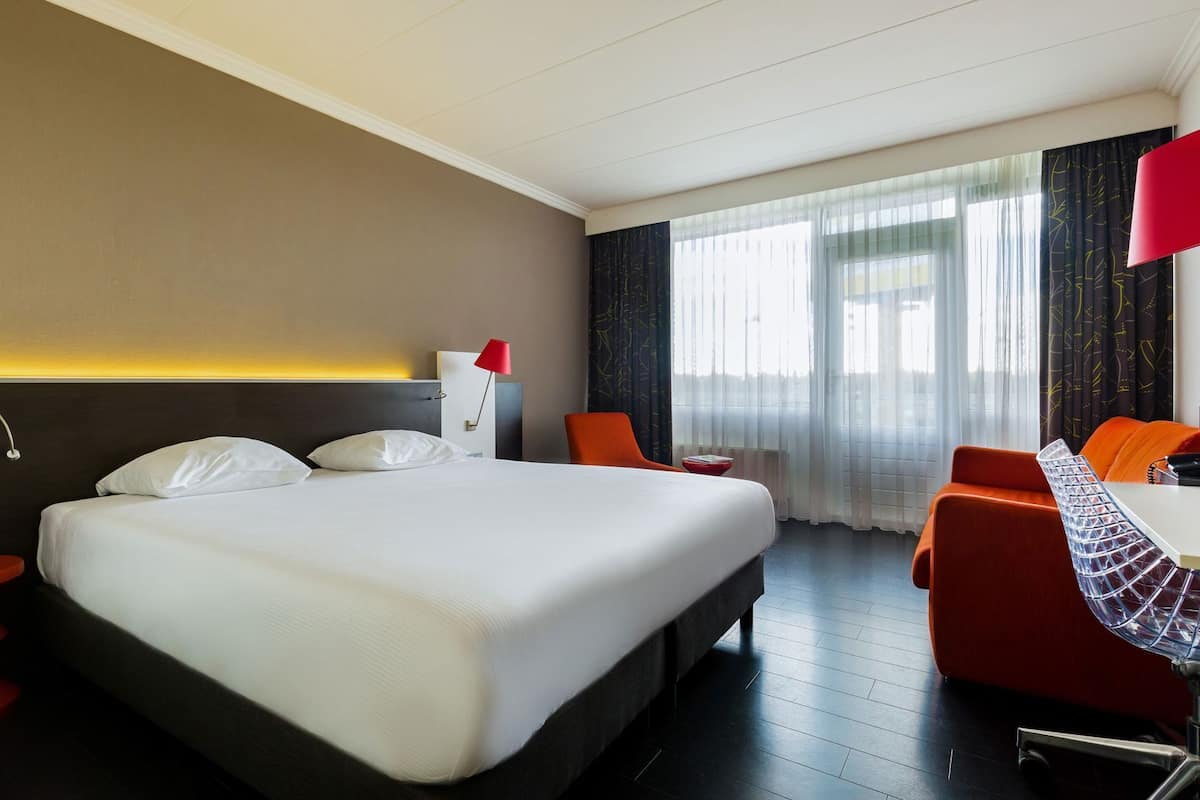 A hotel room with a large bed, a desk with a chair, and a view of the city through the window.