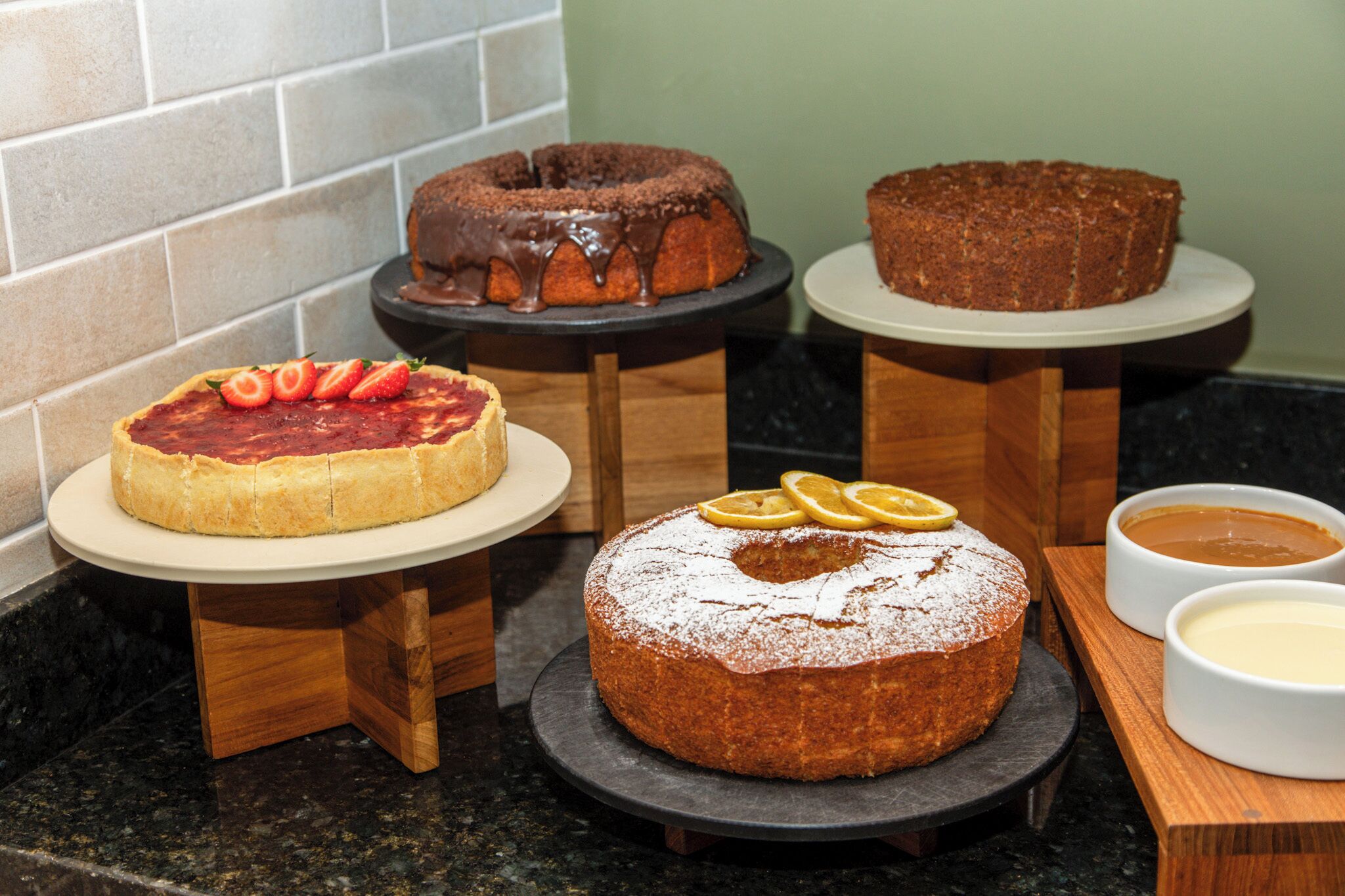 A variety of cakes on wooden stands with a tiled backsplash in the background.