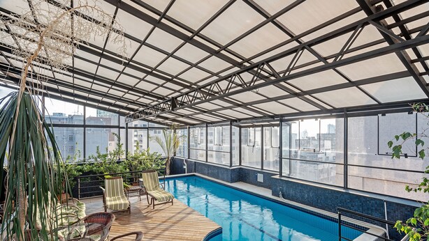 Piscina no terraço com deck de madeira, cadeiras de vime e teto de vidro.