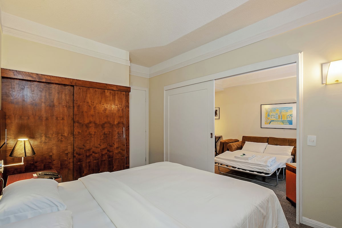 A hotel room with a large bed, a wooden wardrobe, a bedside table with a lamp, and a painting on the wall.