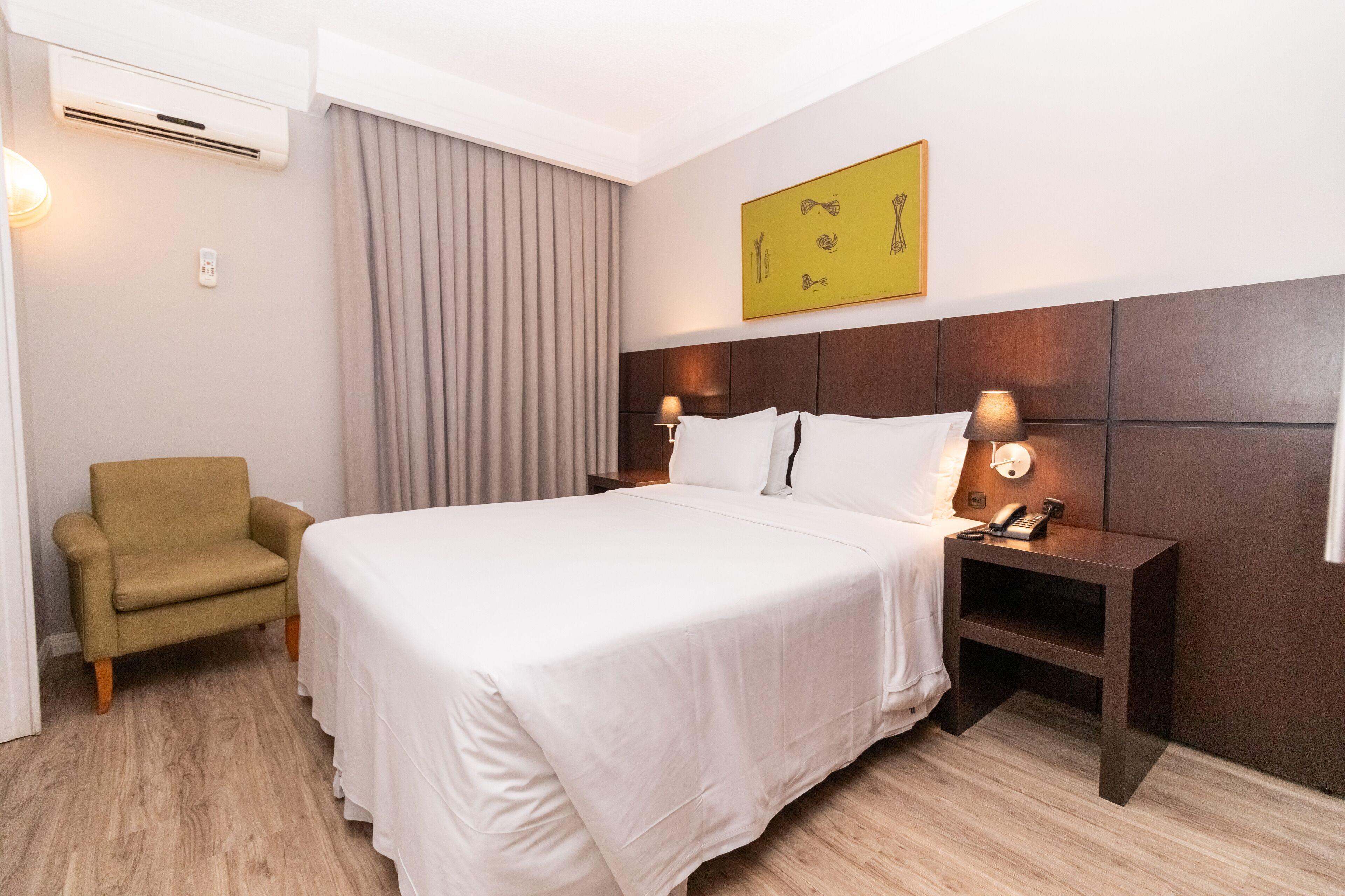 Une chambre d’hôtel avec un grand lit, une table de chevet avec une lampe, une chaise, un appareil de climatisation et un tableau au mur.