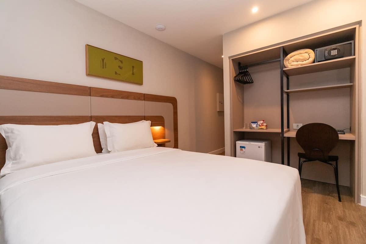A hotel room with a bed, a nightstand, a desk with a chair, a microwave, and a shelf with various items.