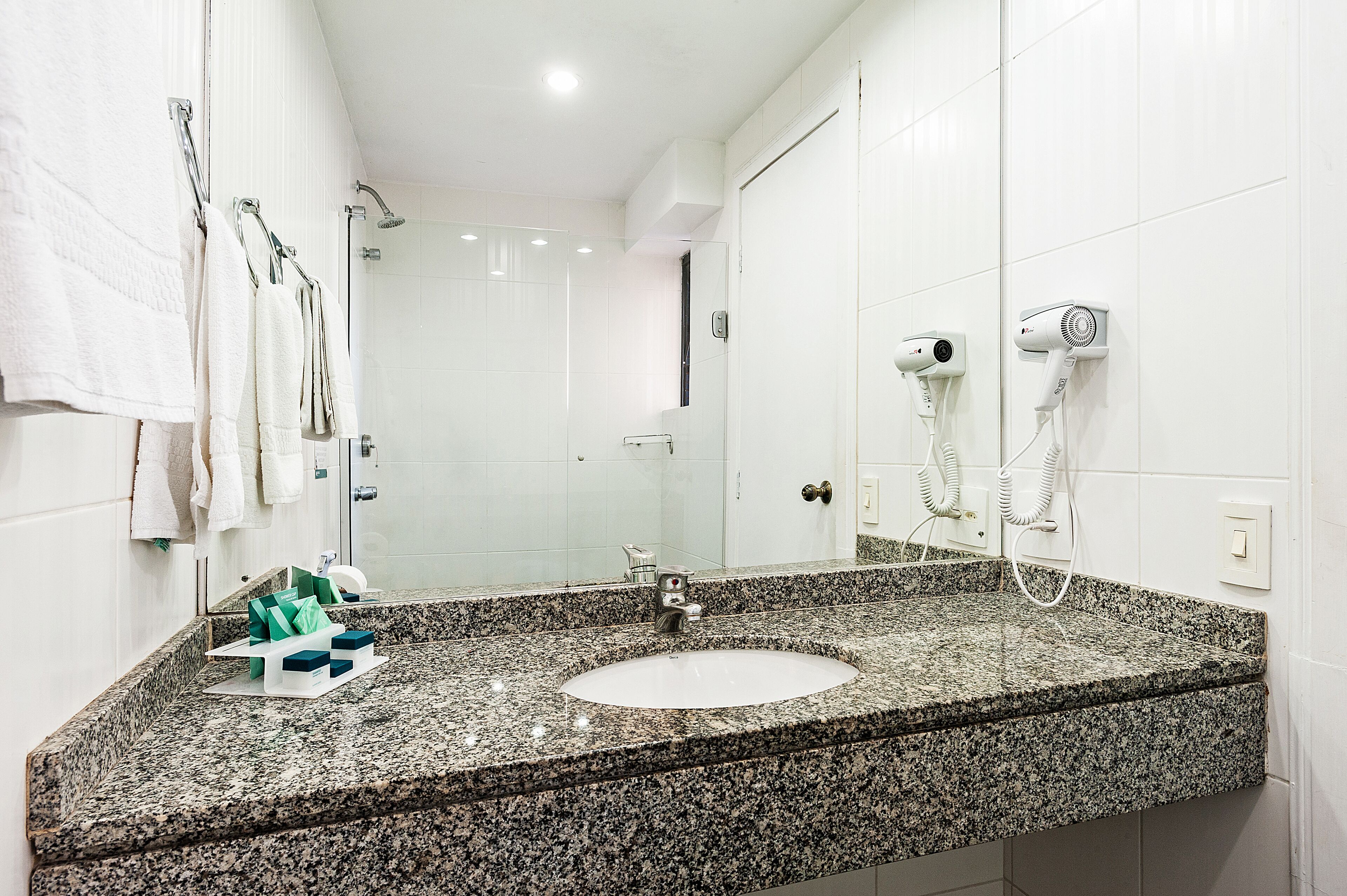 A bathroom with a granite countertop sink, a mirror, a towel rack, a shower, and a hairdryer mounted on the wall.