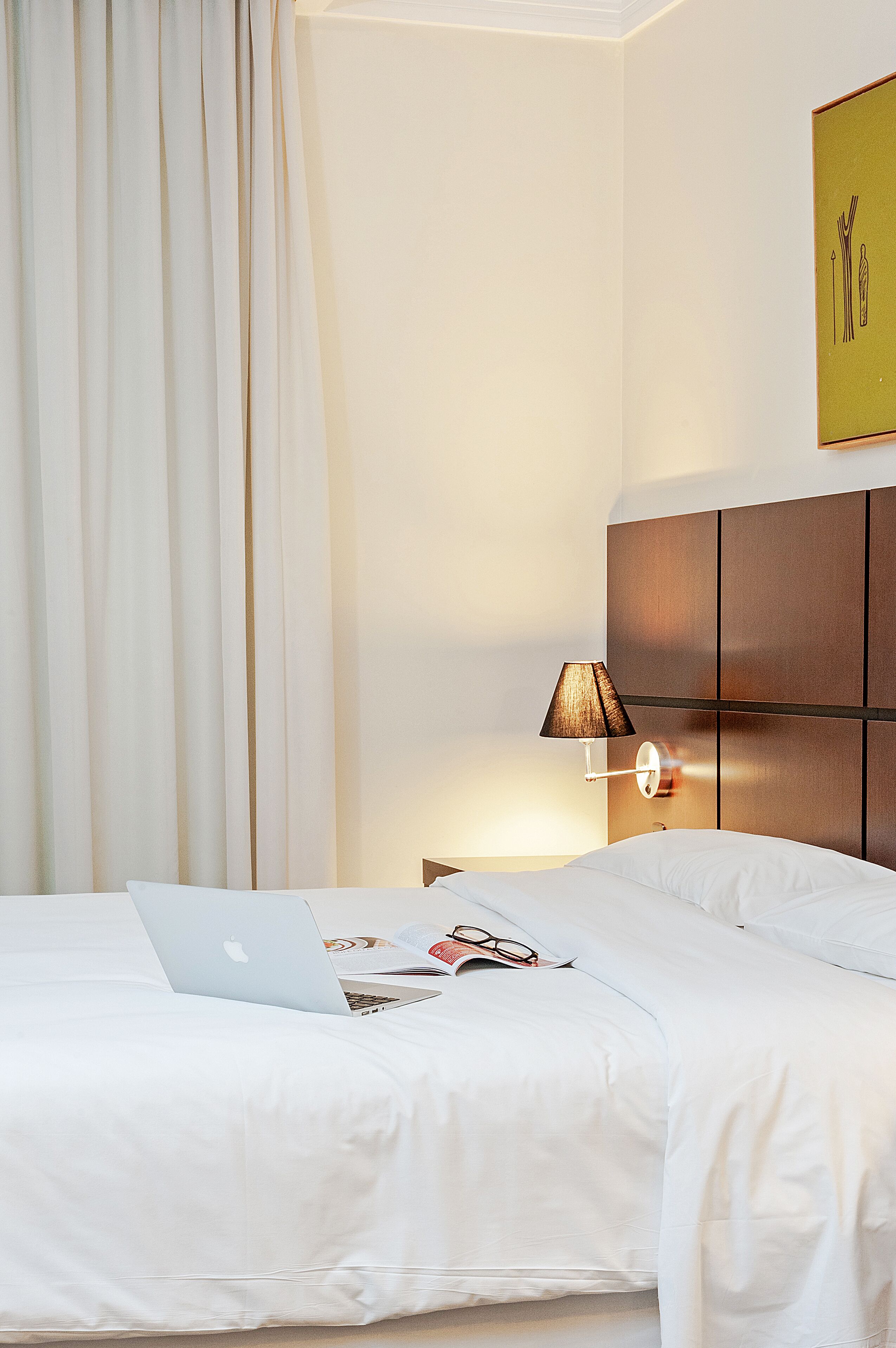 A hotel room with a bed, a laptop, a lamp, and a framed picture on the wall.