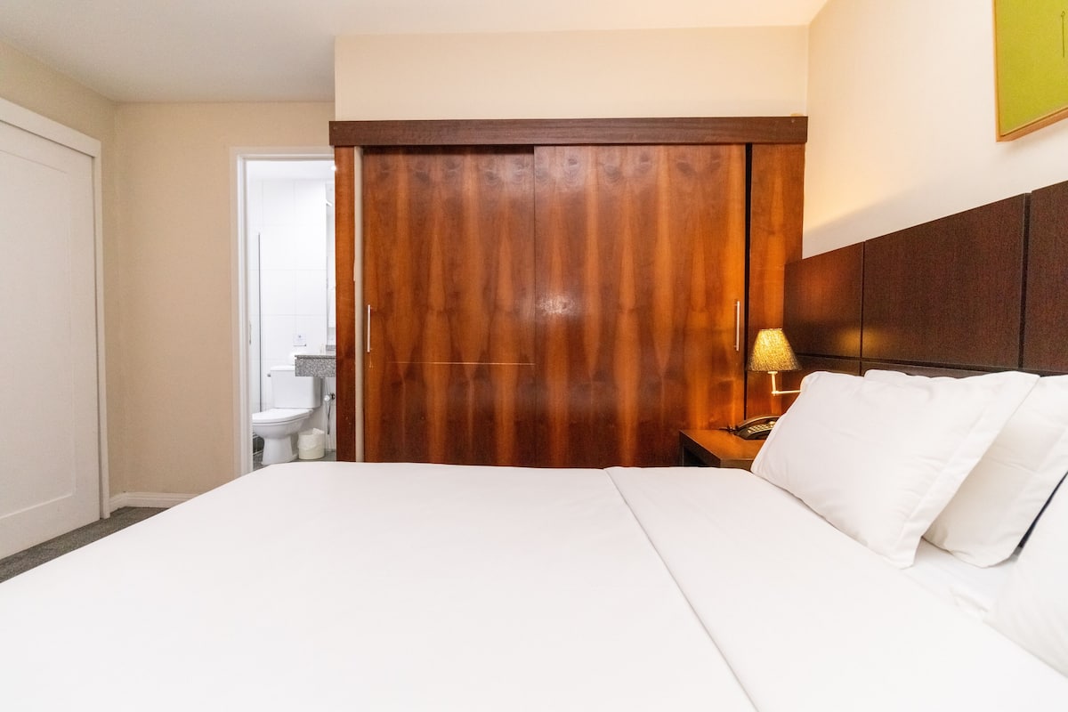 A hotel room with a large bed, a wooden wardrobe, a bedside table with a lamp, and a view of the bathroom.