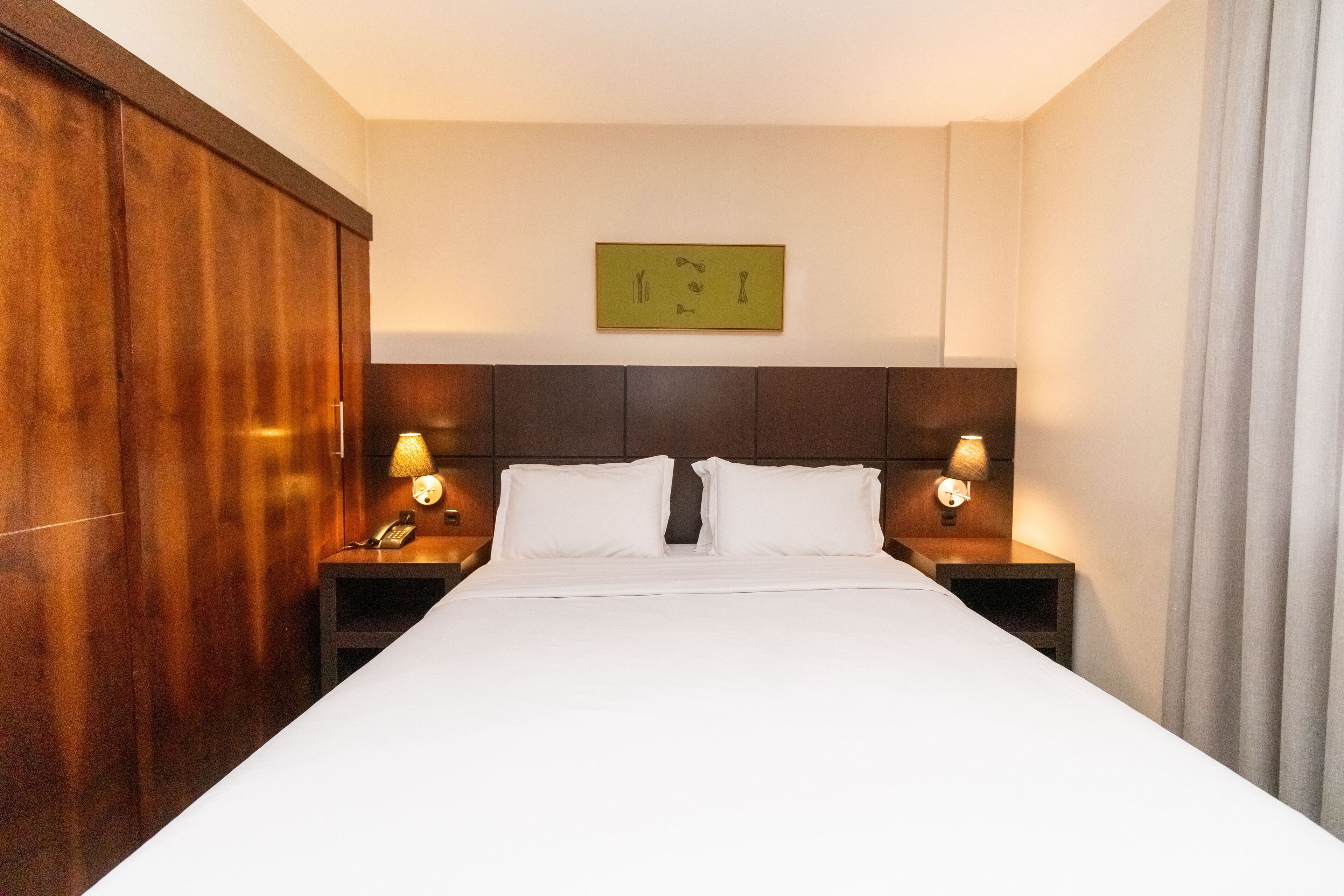 Quarto de hotel com uma cama grande, guarda-roupa de madeira, duas mesas de cabeceira com abajures e uma obra de arte na parede.