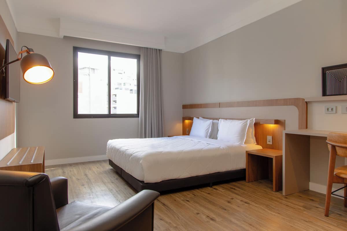 A hotel room with a bed, bedside tables, a desk, a chair, and a window with curtains.