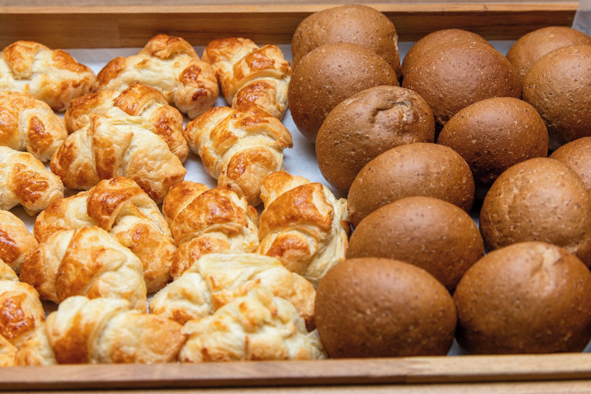 A variety of baked goods, including croissants and round bread rolls, displayed in a wooden tray.