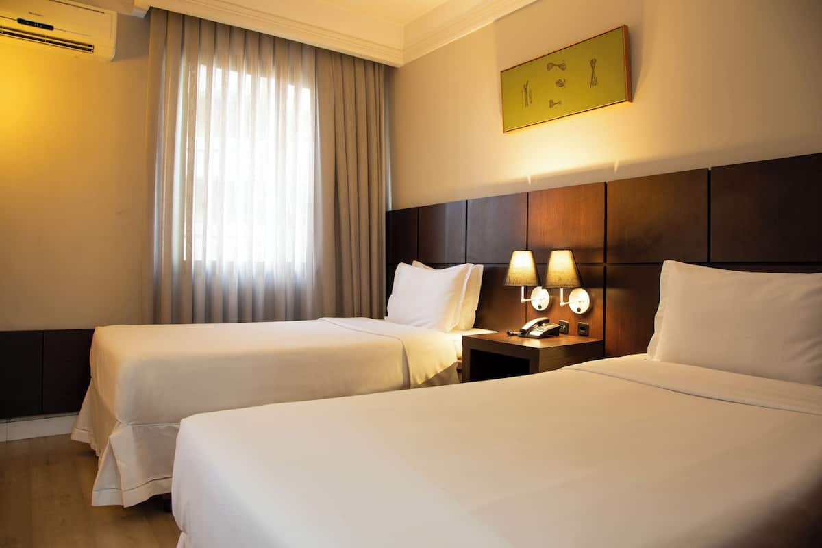 A hotel room with two beds, a nightstand with lamps, a wall-mounted artwork, and a window with curtains.