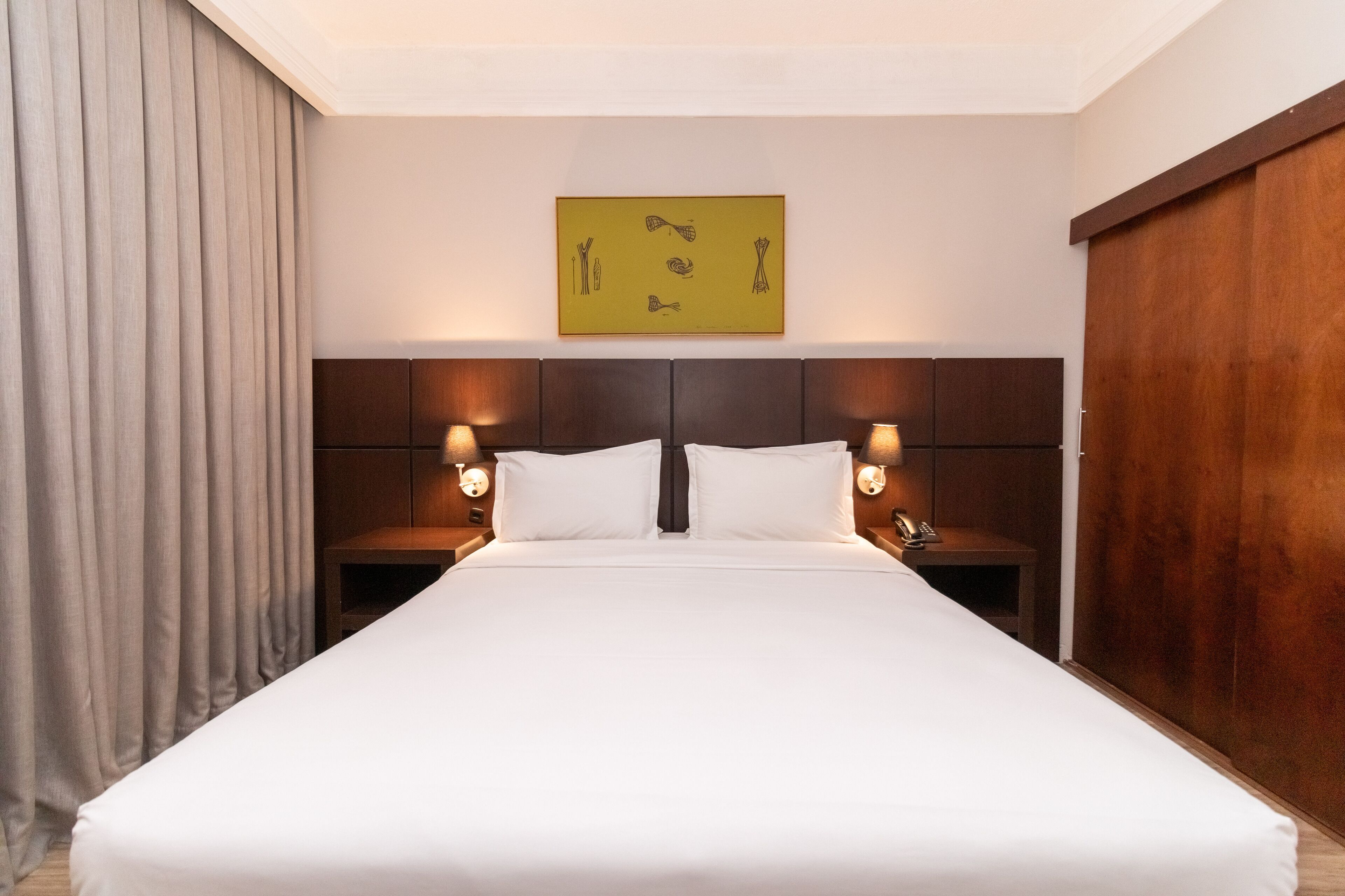 A hotel room with a large bed, wooden headboard, two bedside tables with lamps, a wall-mounted artwork, and a large window with curtains.