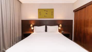 Una habitación de hotel con una cama grande, cabecero de madera, dos mesitas de noche con lámparas, una obra de arte en la pared y un ventanal con cortinas.