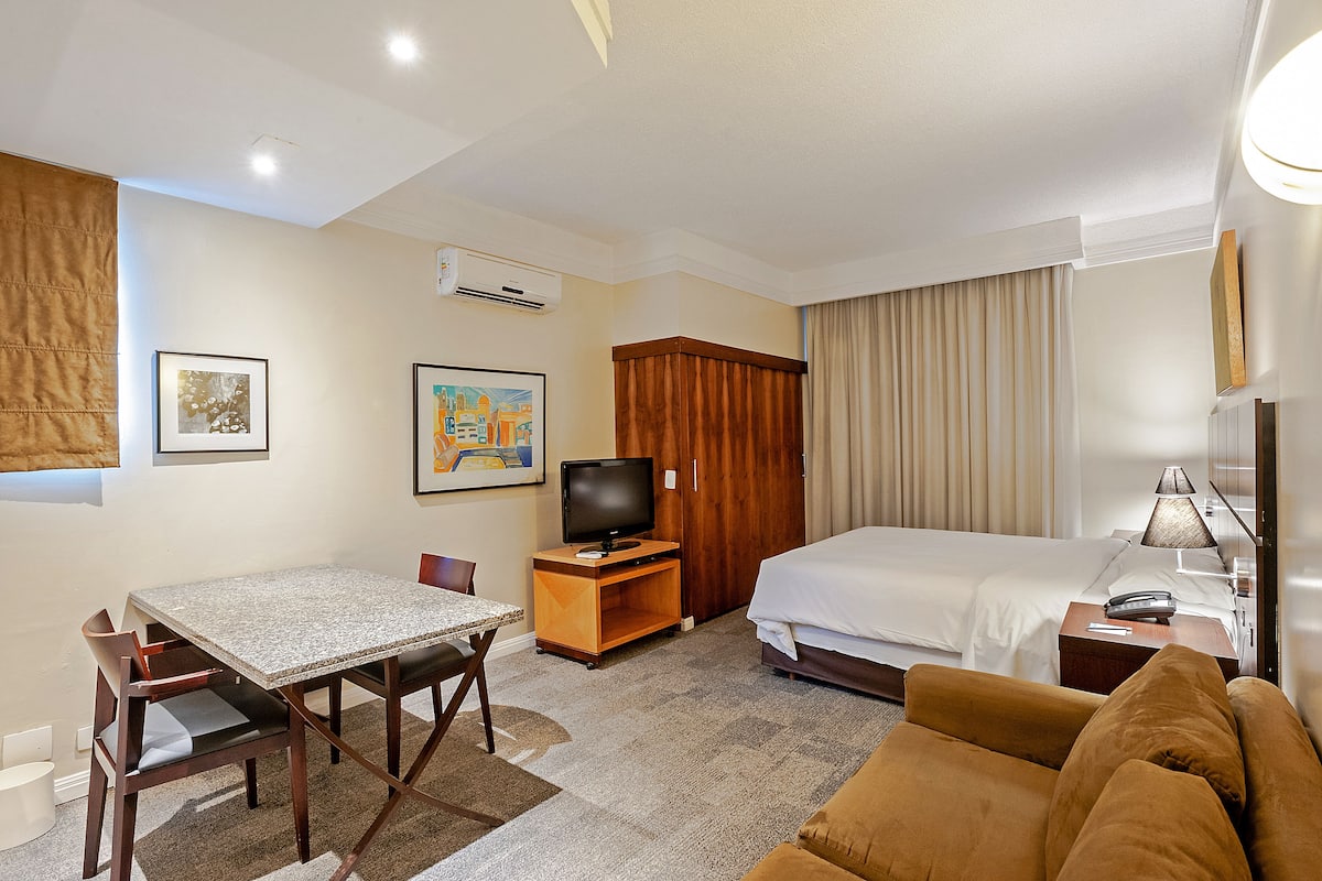 A hotel room with a bed, a TV, a dining table with chairs, a sofa, and a window with curtains.