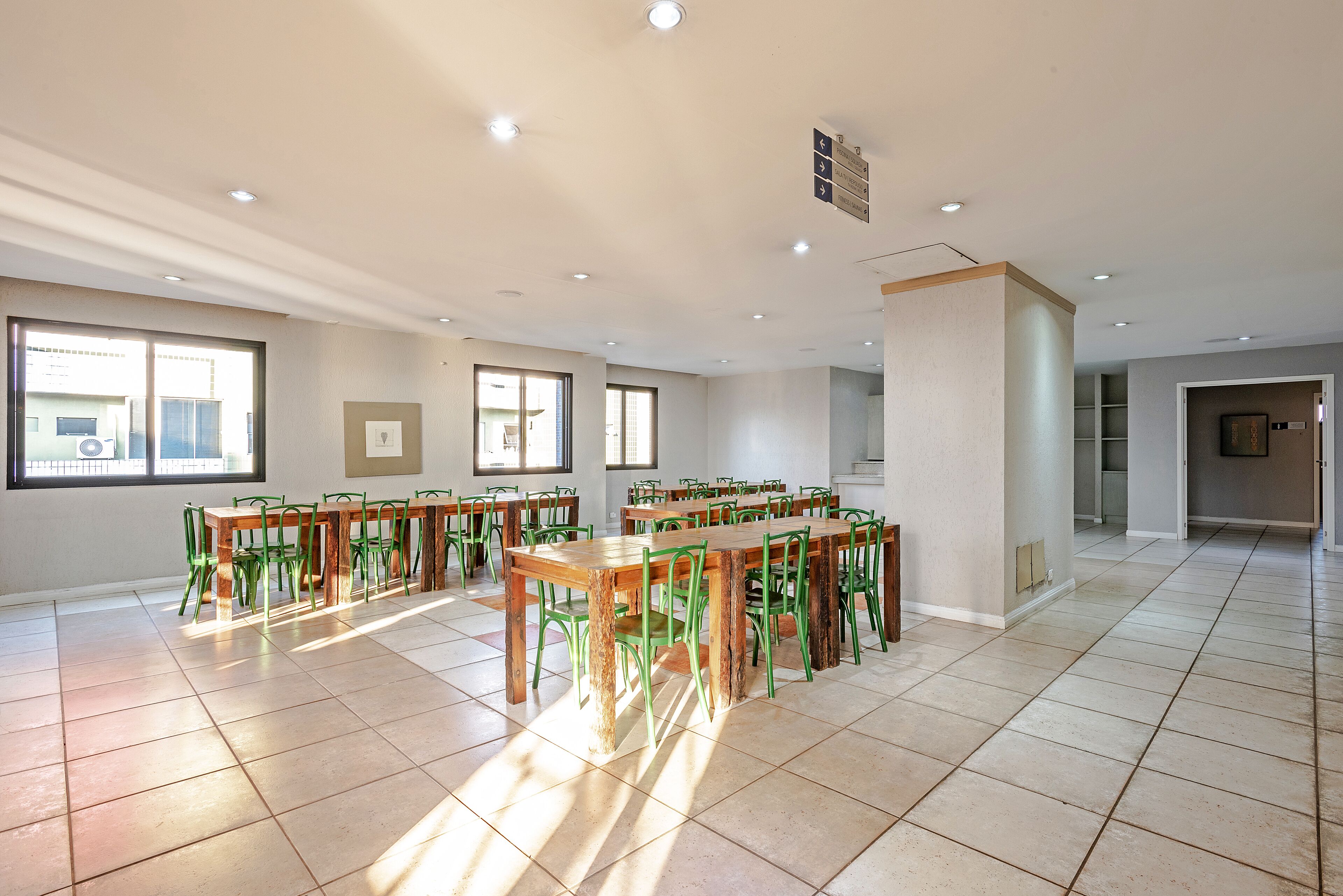 Um quarto amplo com várias mesas de madeira e cadeiras verdes, janelas grandes que permitem entrada de luz natural e piso de azulejos.