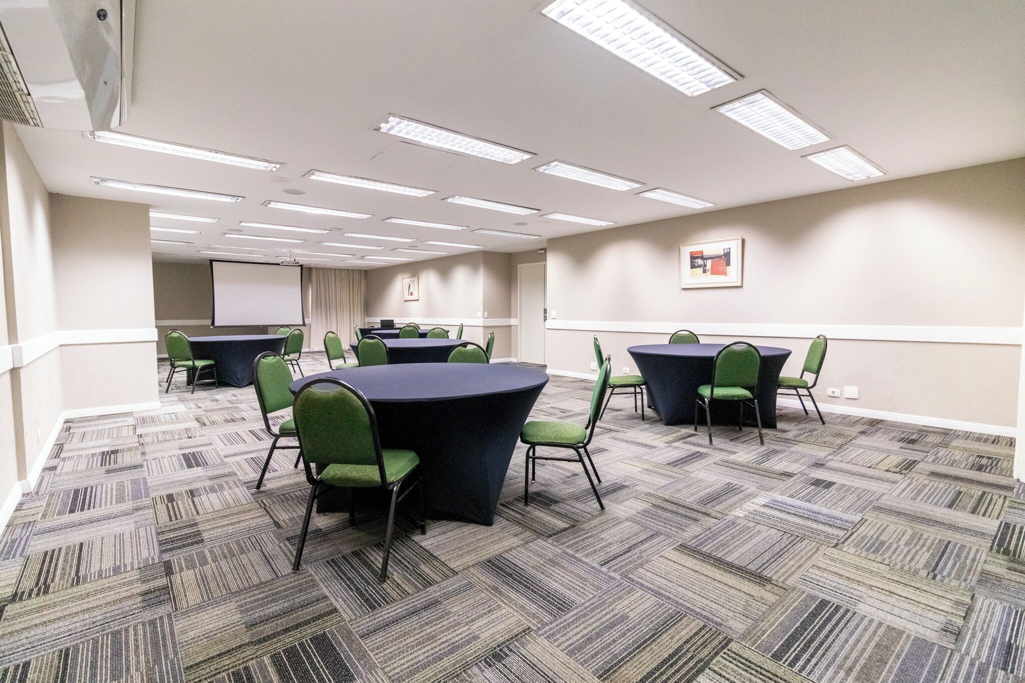 Uma sala de conferências com piso coberto por carpete, cadeiras verdes, uma mesa redonda e uma tela de projeção.