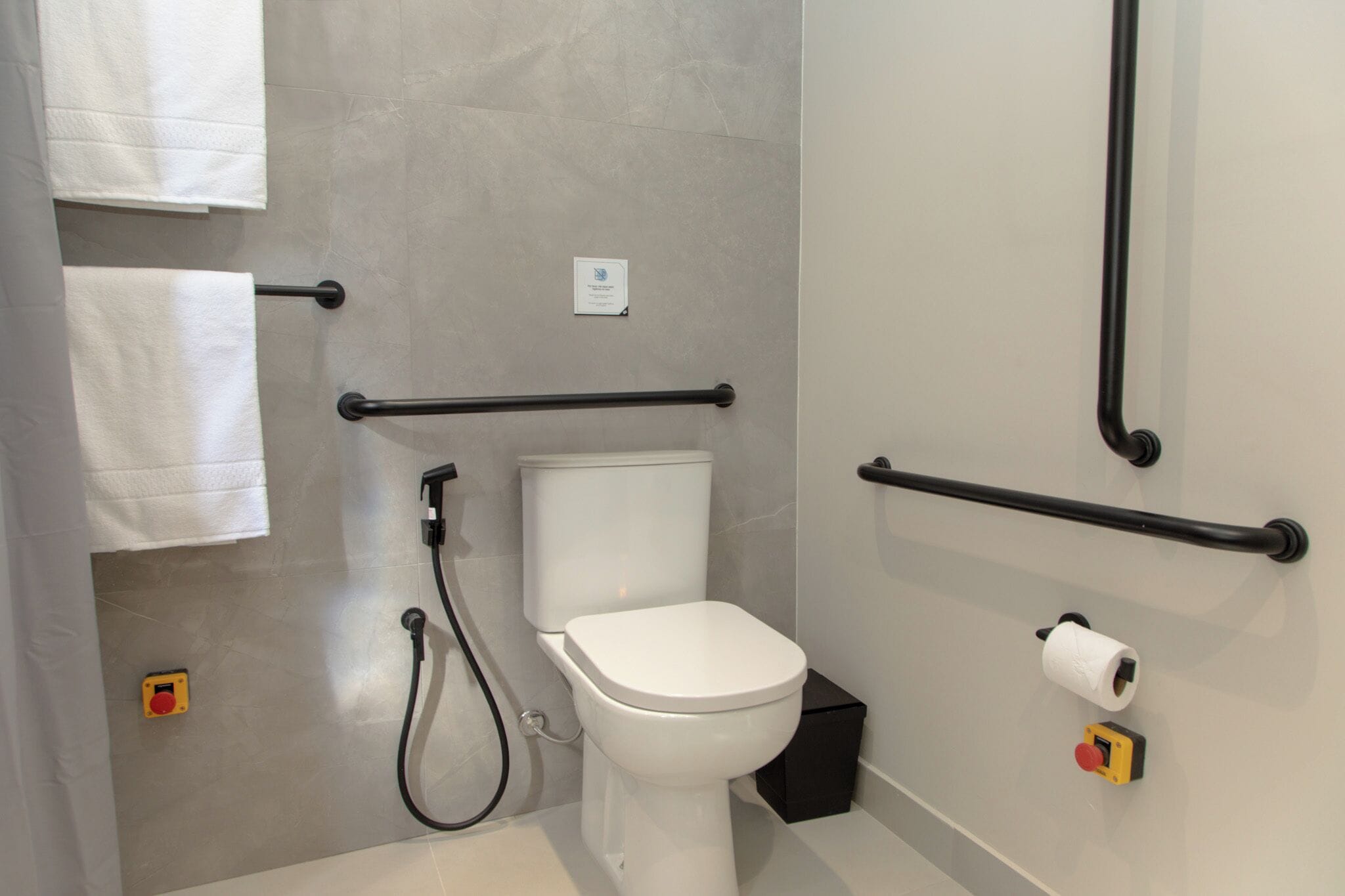 Un baño moderno con inodoro, barras de apoyo, toallero y dispensador de papel higiénico.