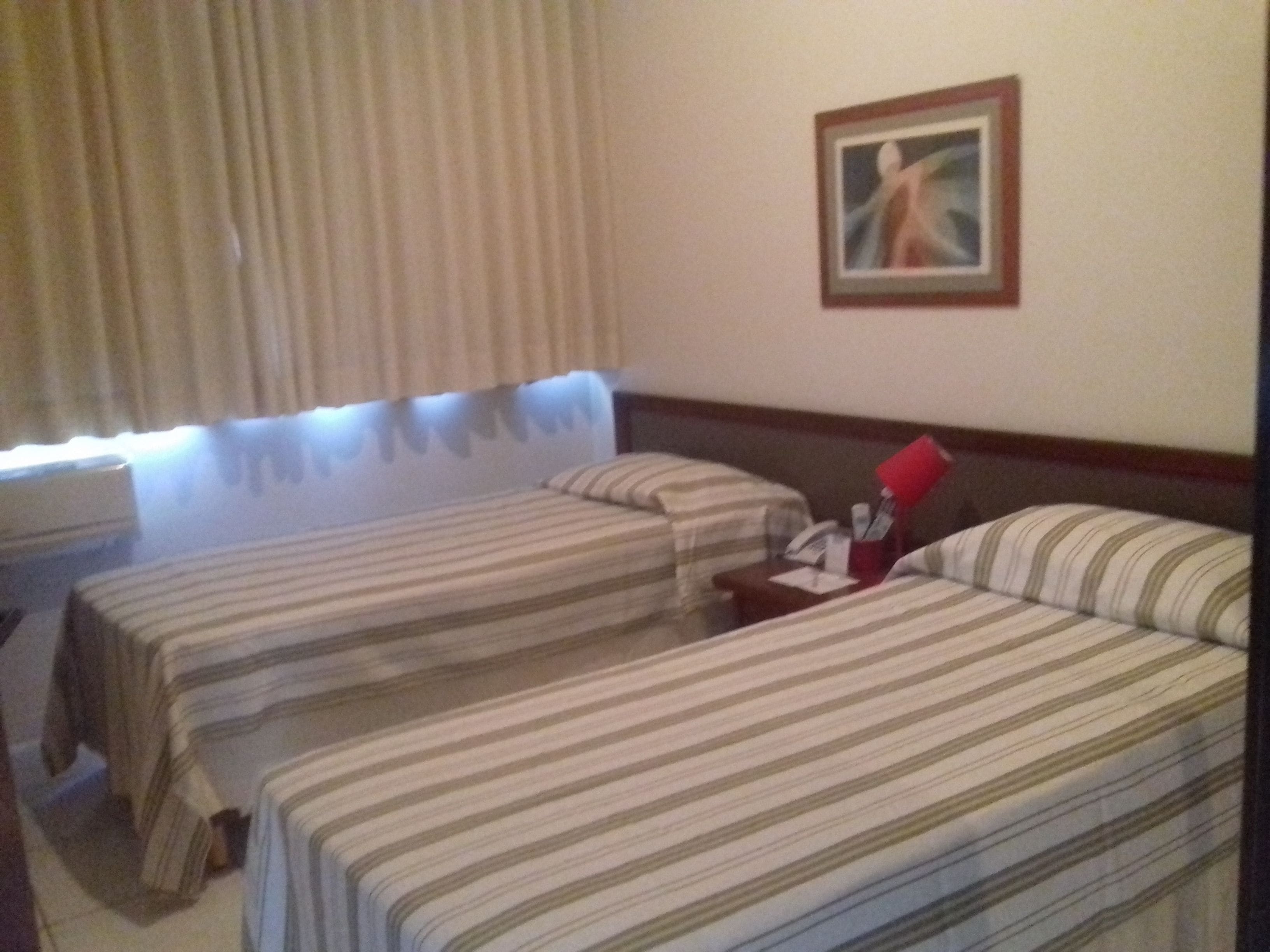 A hotel room with two beds, a nightstand, a painting on the wall, and a window with curtains.