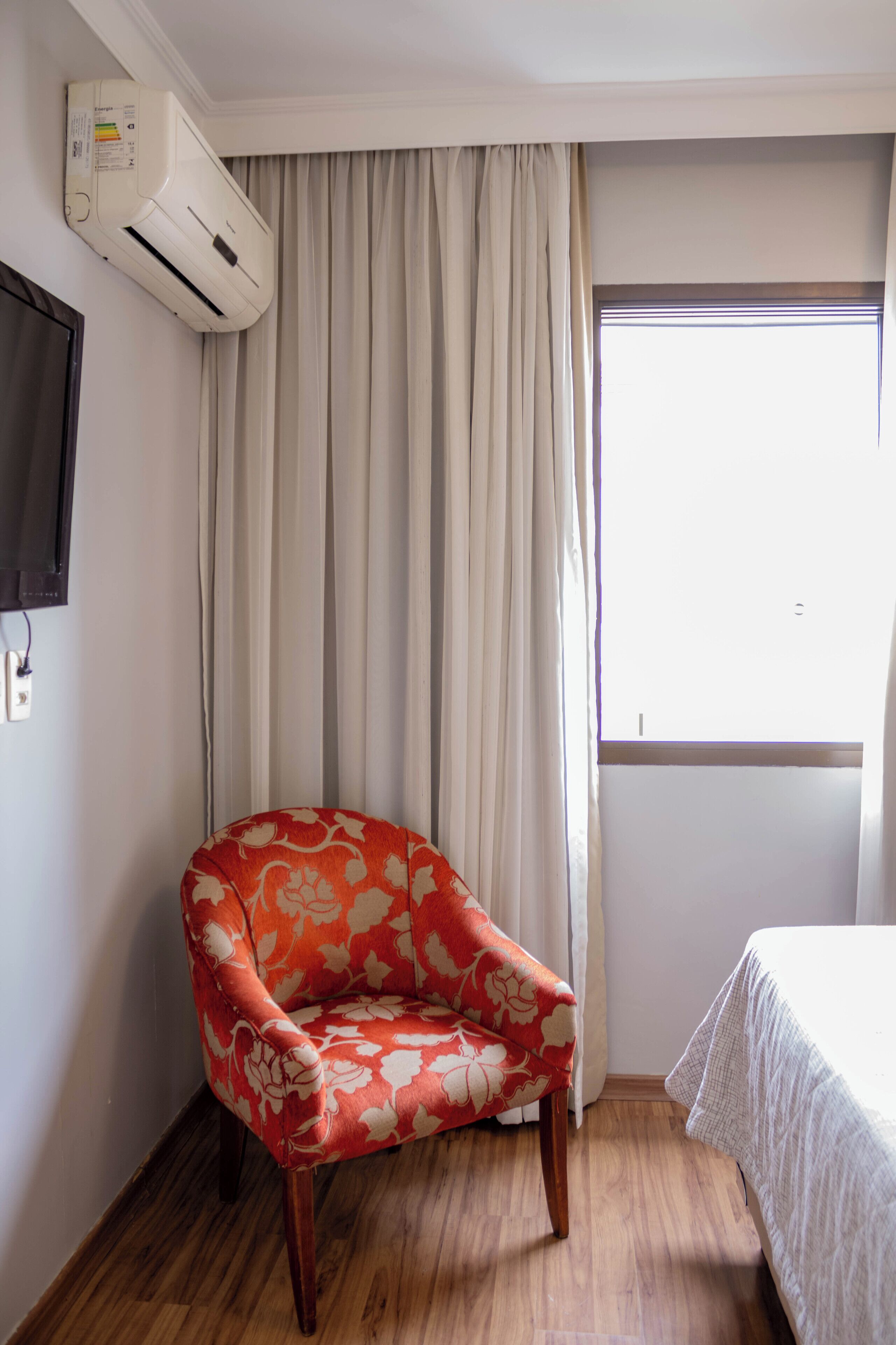 A room with a wooden floor, a red floral armchair, a bed with a white bedspread, a window with beige curtains, and an air conditioning unit on the wall.