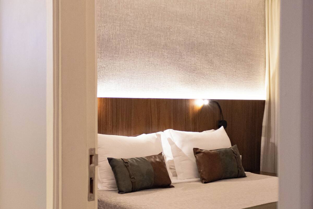Una habitación de hotel con una cama bien hecha, un cabecero con una lámpara y una ventana con cortinas.