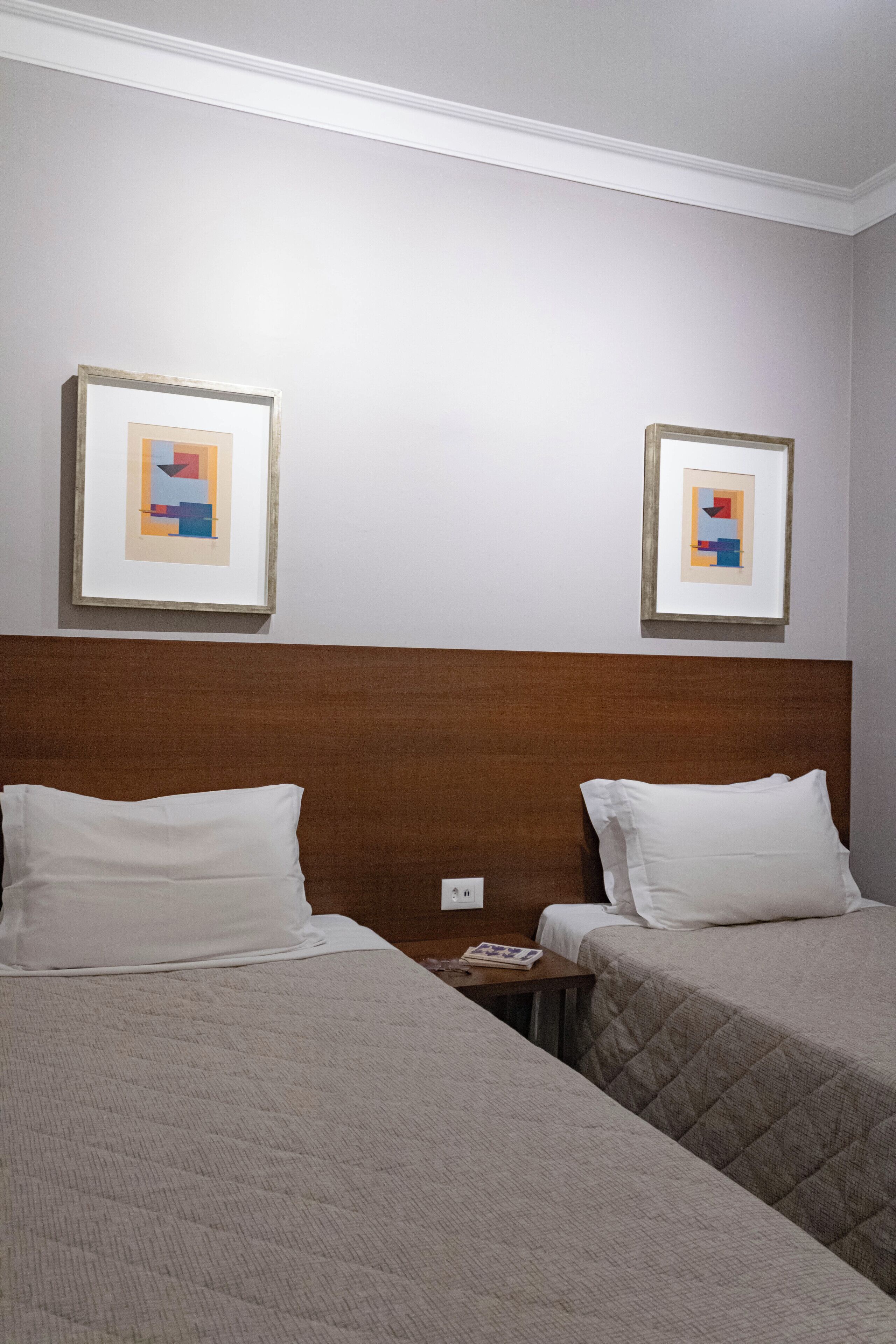 A hotel room with two beds, a wooden headboard, and two framed abstract paintings on the wall.