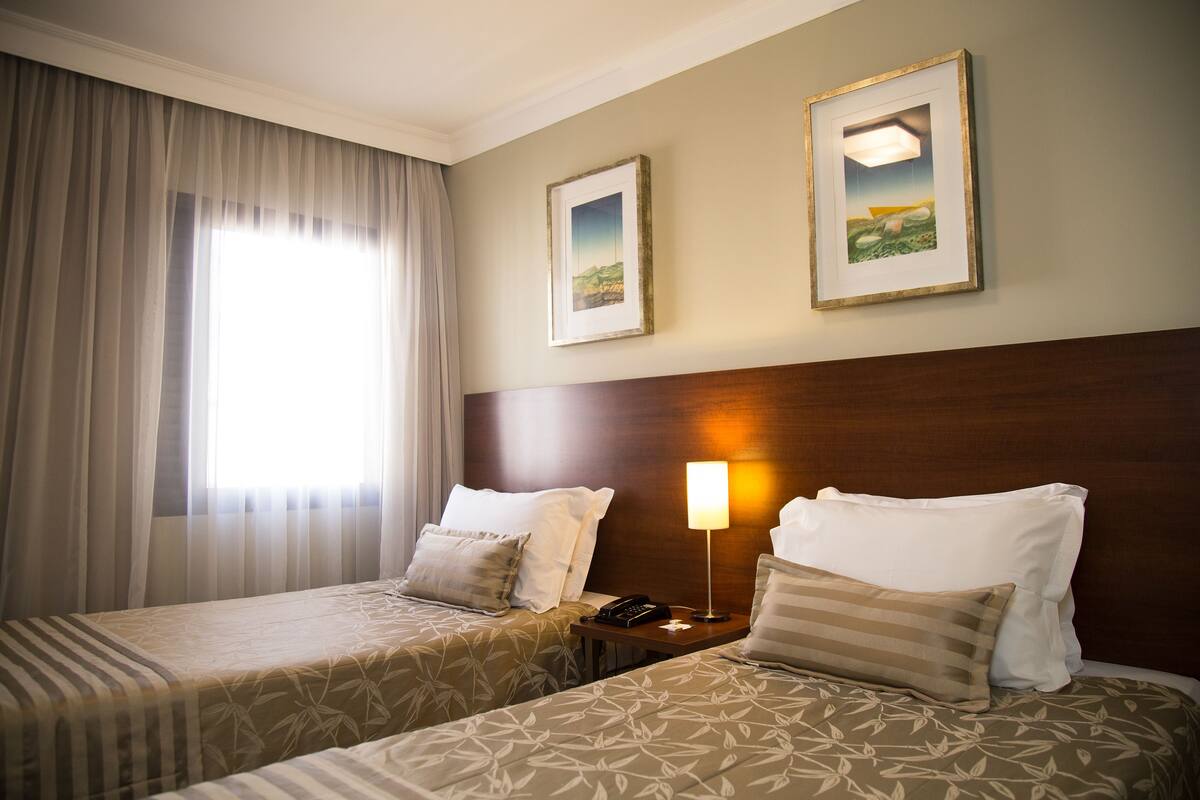 Habitación de hotel con dos camas, una mesita de noche con lámpara y dos cuadros en la pared.