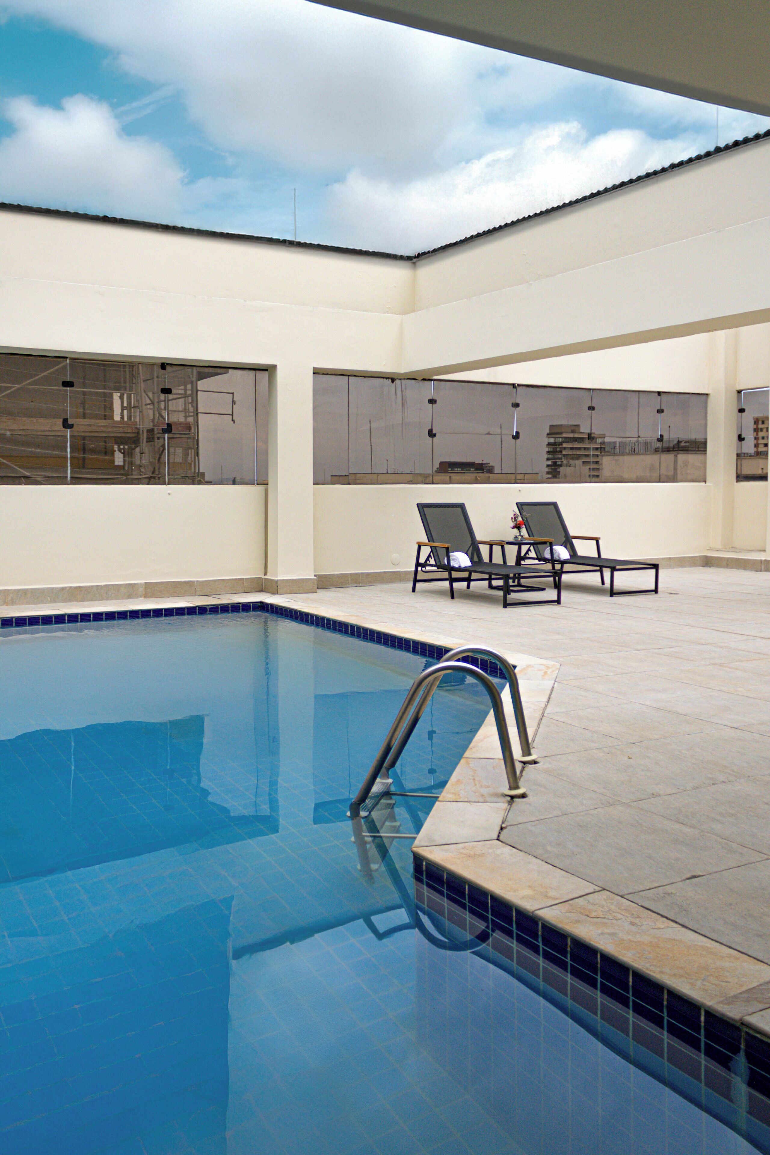 A swimming pool with a clear blue water, surrounded by a tiled deck. There are two lounge chairs and a small table with a flower vase.