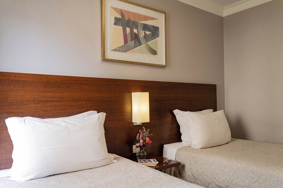 Habitación de hotel con dos camas, cabecero de madera, mesita de noche con lámpara y un cuadro abstracto enmarcado en la pared.