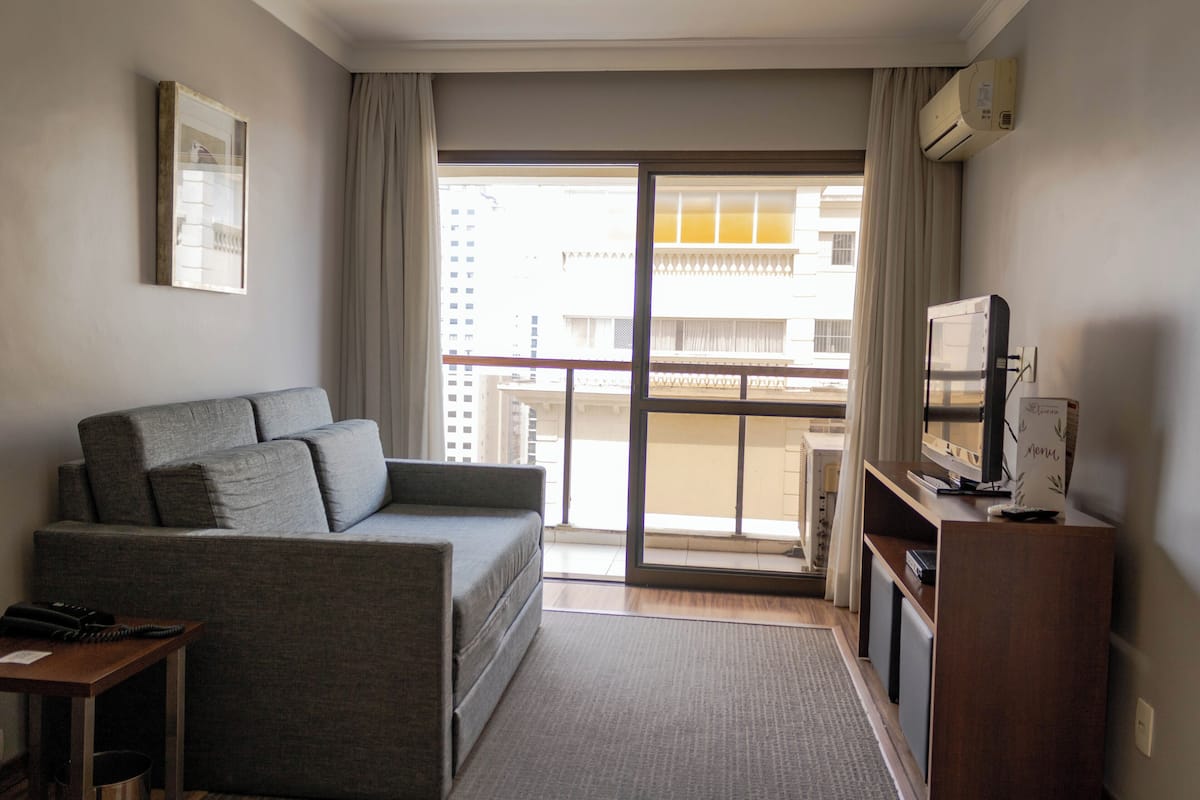 Una habitación de hotel moderna con un sofá gris, una pequeña mesa de madera, un televisor y un balcón con vistas a edificios.