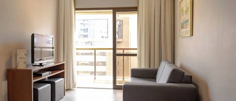 Una sala moderna con un sofá gris, un televisor sobre un mueble de madera y un balcón con cortinas.