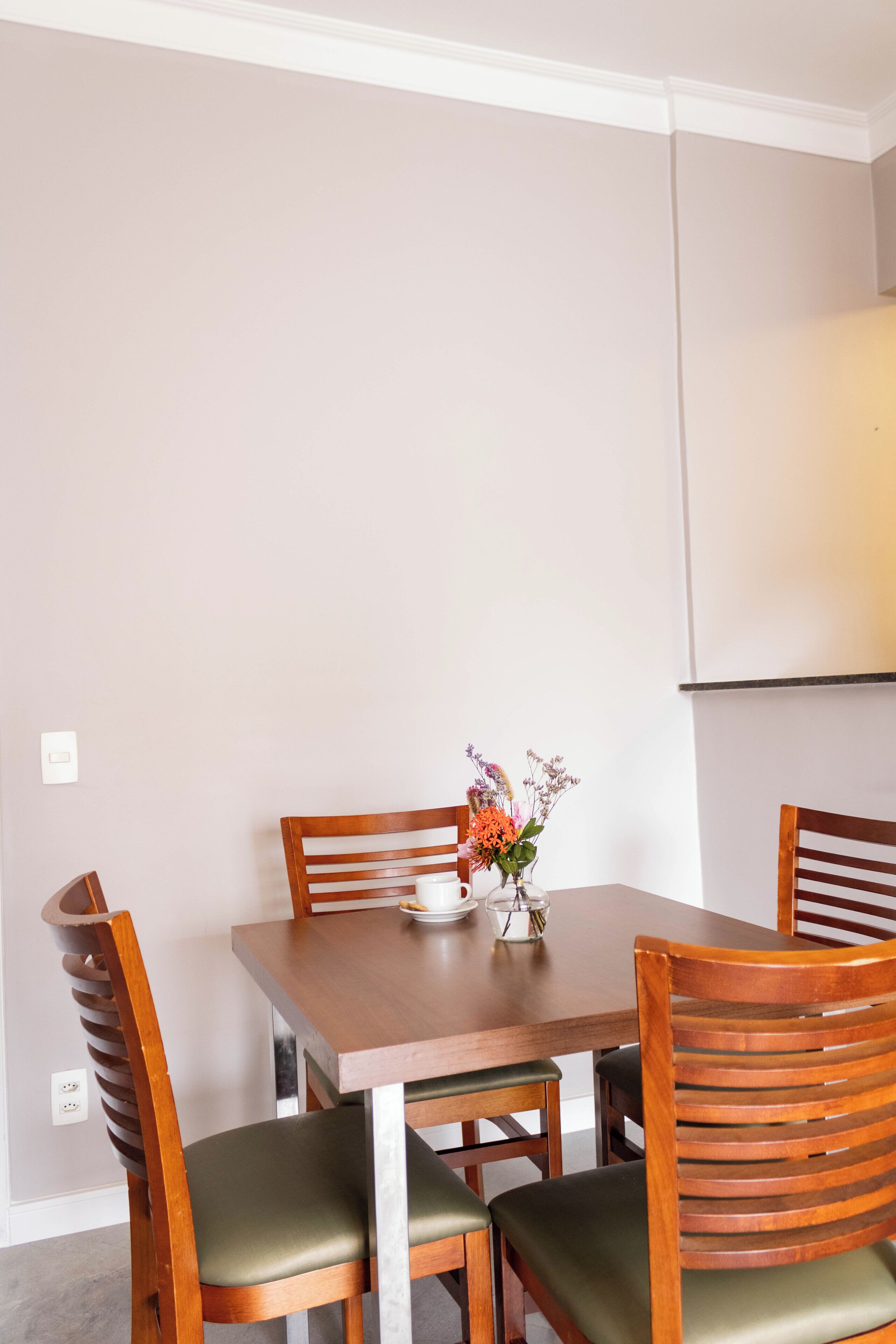A dining area with a wooden table and four chairs, a vase with flowers, and a light switch on the wall.