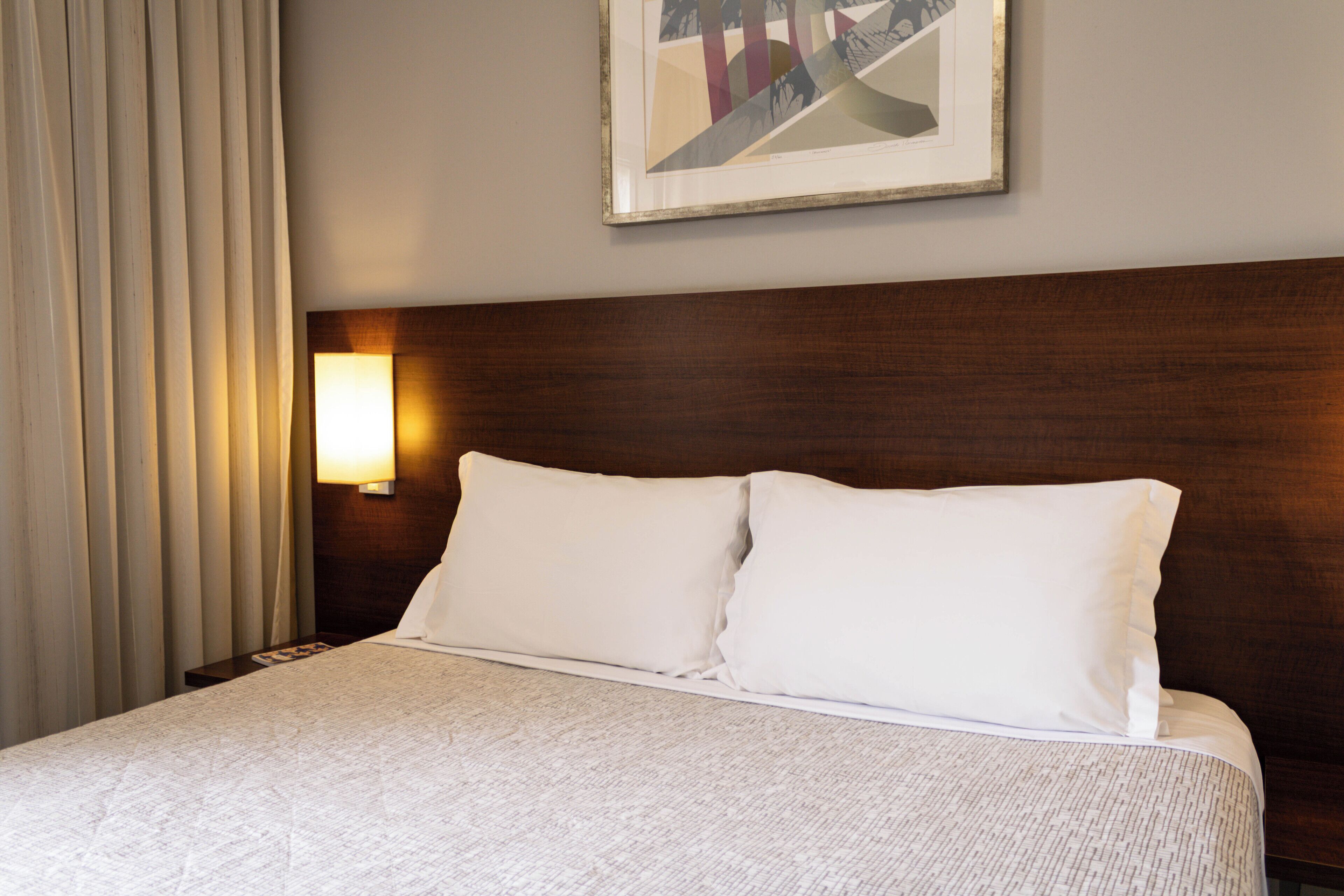 A hotel room with a large bed, a headboard, a wall-mounted lamp, and a framed picture on the wall.
