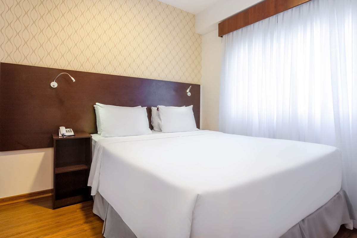 A hotel room with a large bed, a bedside table with a phone, a headboard, and a window with curtains.