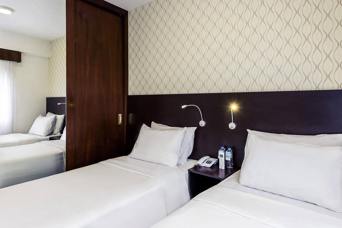 A hotel room with two single beds, a wooden door, a patterned wallpaper, and a bedside table with a phone and water bottles.