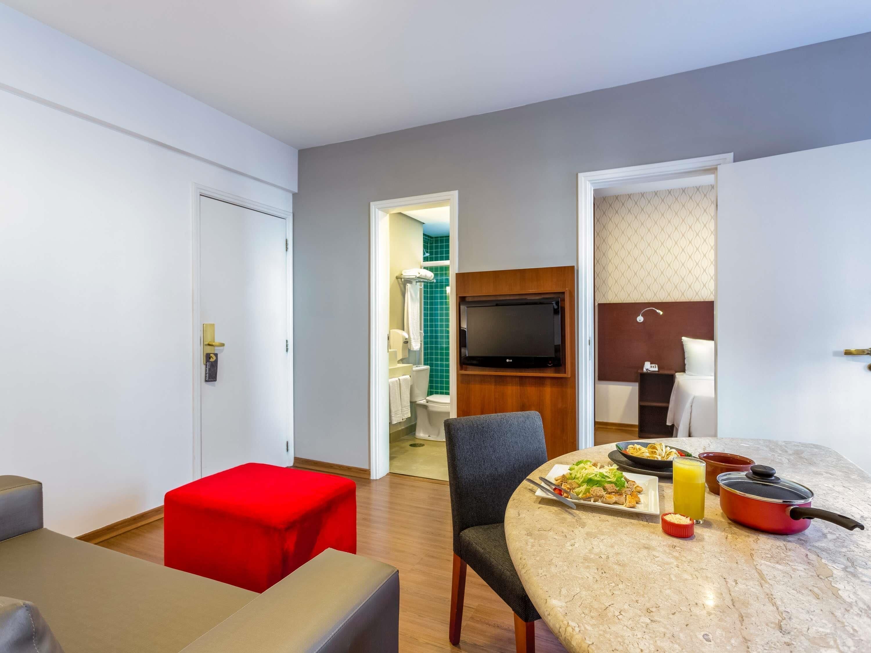 Camera d'hotel moderna con zona pranzo, TV fissata su un pannello di legno e bagno visibile attraverso una porta aperta.