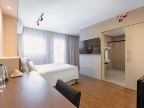 Une chambre d’hôtel comprenant un lit, un bureau avec une chaise, un téléviseur fixé au mur, un espace douche et une porte avec une poignée.