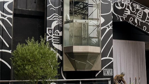 Un área de estar moderna al aire libre con dos sillas azules y una mesa redonda, rodeada de follaje y un ascensor de vidrio.